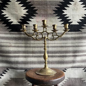 May include: A vintage brass candelabra with five candle holders. The candelabra is on a small wooden stool. The background features a black, white, and gray patterned textile.