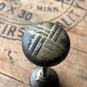 May include: Antique brass doorknob with a weathered finish. The round knob features a crosshatch design and a dark, aged patina. The stem and base are also made of brass, showing signs of age and use. The background is a wooden surface.