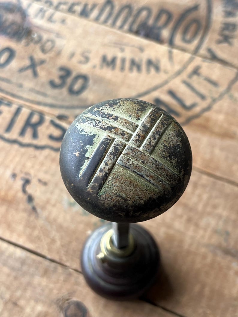 May include: Antique brass doorknob with a weathered finish. The round knob features a crosshatch design and a dark, aged patina. The stem and base are also made of brass, showing signs of age and use. The background is a wooden surface.