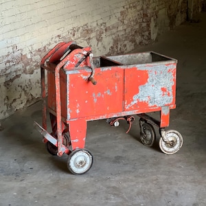 May include: An antique, red metal cart with a rectangular bin and four wheels. The cart has a hand crank and a handle. The paint is chipped, revealing the metal underneath. The wheels are white and gray.