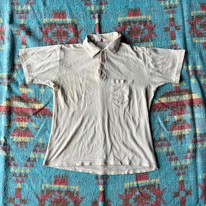 May include: A light beige short-sleeved polo shirt with a button-up collar and a single pocket on the left chest.