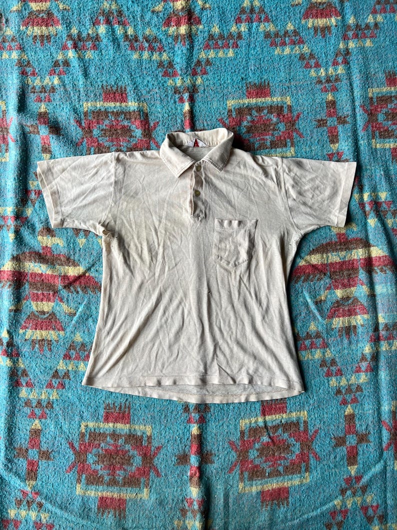 May include: A light beige short-sleeved polo shirt with a button-up collar and a single pocket on the left chest.