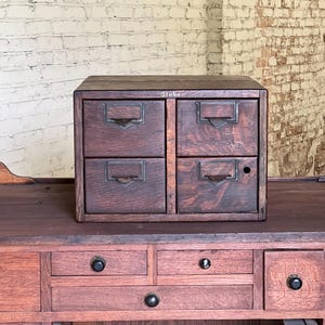 May include: A small, dark wooden cabinet with four drawers, each featuring a metal pull and label holder. The word "Globe" is printed on the top. The cabinet sits on a larger wooden desk, against a white brick wall.
