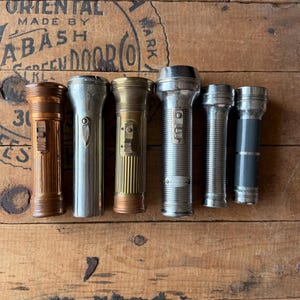 May include: A collection of six vintage metal flashlights arranged in a row. The flashlights vary in color, including copper, brass, and silver tones, and feature different textures, ribbed barrels, and sliding power switches on their bodies.