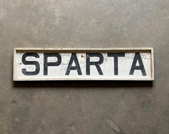 May include: A white wooden sign with black painted letters spelling "SPARTA".