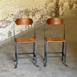 May include: Two vintage chairs with wooden seats and backs, and dark metal frames. The chairs have a simple, industrial design, with curved legs and a minimalist aesthetic. The wood has a warm, natural tone.