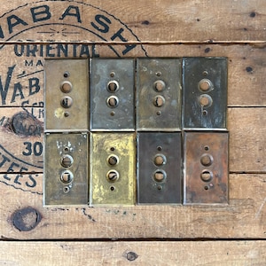 May include: Eight vintage brass and metal light switch covers with two circular holes for the switches. The covers have a rectangular shape and are arranged on a wooden surface. The covers have a weathered, aged appearance.