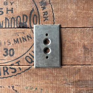 May include: A silver metal switch plate with two round holes and a rectangular shape. The plate is mounted on a wooden surface.