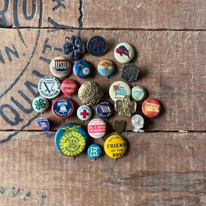 May include: A collection of vintage enamel pins featuring various designs, including a bee, a buffalo, a cross, a heart, a star, a flag, and a 4-H clover. The pins are arranged in a circle on a wooden surface.