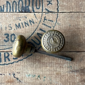 Puede incluir: Dos pomos de puerta antiguos de latón. Uno es redondo con un emblema detallado, el otro tiene una forma redondeada más tradicional. Ambos pomos tienen una varilla roscada. Los pomos están sobre una superficie de madera envejecida.