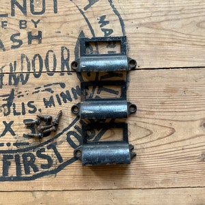 May include: Three black metal drawer pulls with rounded edges and a rectangular opening. The pulls are arranged vertically on a wooden surface with a stamp that reads "First National Bank of St. Louis, Minn."