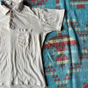 May include: A light beige short-sleeved polo shirt with a single button placket and a small pocket on the left chest.