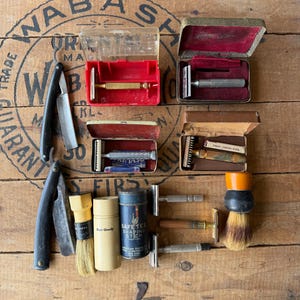 May include: An assortment of vintage shaving tools, including razors, brushes, and shaving sticks, arranged on a wooden surface. The items are in various conditions, with some in cases. The image features a weathered aesthetic, suggesting age and use.