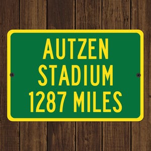May include: A green and yellow sign with the text "AUTZEN STADIUM 1287 MILES". The sign has a yellow border and is mounted on a wooden background. The text is in a bold, sans-serif font.