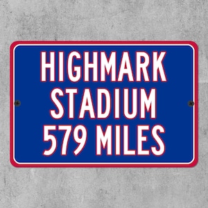 Può includere: Un cartello rettangolare con sfondo blu e testo bianco: "HIGHMARK STADIUM 931 chilometri". Il cartello ha un bordo rosso ed è montato su un muro grigio.