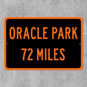Op de afbeelding: Zwart rechthoekig bord met oranje tekst "ORACLE PARK" en "72 MILES". Het bord heeft een oranje rand en is bevestigd aan een grijze betonnen muur. Twee schroeven zijn zichtbaar.