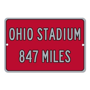 Puede incluir: Un letrero rectangular rojo con las palabras "OHIO STADIUM" en letras grises grandes en la parte superior y "847 MILES" en letras grises más pequeñas debajo. El letrero tiene un borde gris y dos agujeros pequeños.
