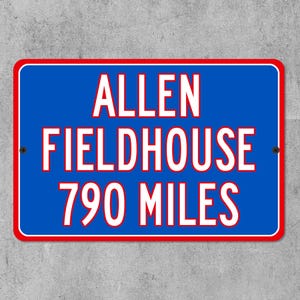May include: A rectangular blue sign with a red border and white text that reads "ALLEN FIELDHOUSE 790 MILES". The sign is mounted on a gray concrete wall.