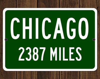 Puede incluir: Un letrero rectangular verde con texto blanco que dice "CHICAGO" encima de "2387 MILES". El letrero tiene un borde blanco y está montado sobre un fondo de madera.
