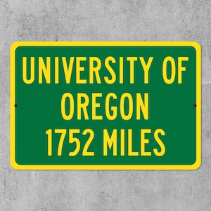 May include: A rectangular green sign with yellow text and a yellow border. The sign reads "UNIVERSITY OF OREGON 1752 MILES". The sign is mounted on a grey concrete wall.
