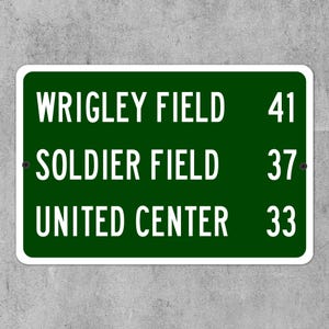May include: A rectangular green sign with white text. The sign lists "WRIGLEY FIELD 41", "SOLDIER FIELD 37", and "UNITED CENTER 33". The sign has a white border and is mounted on a gray surface.