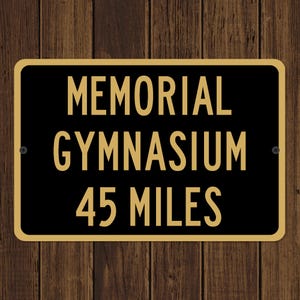 May include: Black and gold rectangular sign with the words "MEMORIAL GYMNASIUM 45 MILES" in gold lettering. The sign is mounted on a wooden background.