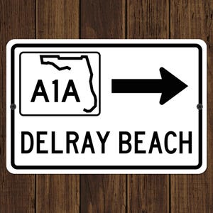 May include: A white rectangular sign with black text and a black border. The sign reads "A1A" with a Florida outline, an arrow pointing right, and "DELRAY BEACH" below. The sign is mounted on a wooden surface.