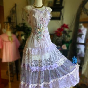 Könnte beinhalten: Vintage lavendelfarbenes Kleid auf einer Schaufenpuppe. Das Kleid hat ein floral besticktes Oberteil, Rüschenspitze und einen mehrschichtigen Rock. Das Kleid hat eine zarte, romantische Ästhetik mit fließender Silhouette und einem Hauch von Laune.