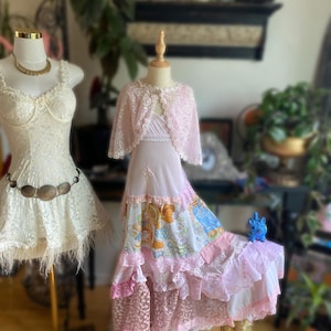 May include: Two mannequins display vintage dresses. One is a white lace dress with a gold necklace and belt. The other is a pink dress with a lace bolero. The pink dress has a patchwork skirt with cartoon characters and lace trim.