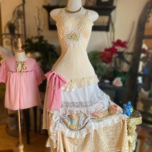 May include: A vintage-style dress form displays a cream-colored slip with a lace-trimmed skirt. A pink bow accents the waist. A smaller pink dress is on a separate form. The overall aesthetic is vintage and feminine.