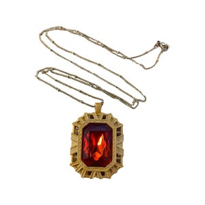 Op de afbeelding: Een goudkleurige ketting met een grote, rechthoekige, rode edelsteen hanger. De hanger is gezet in een decoratief frame. De ketting is delicaat en hangt tegen een witte achtergrond.
