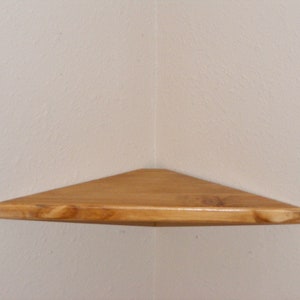 May include: A wooden corner shelf with a triangular shape. The shelf is made of light-colored wood and is mounted on a neutral-colored wall. The shelf is designed to fit into a corner, maximizing space.