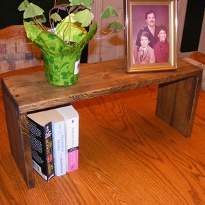 May include: A wooden shelf unit with a dark brown stain. The shelf is 18 inches long and 6 inches wide. It has a small plant with green leaves in a green pot on the top shelf. There are three books on the bottom shelf. A framed photo of three people is on the top shelf.