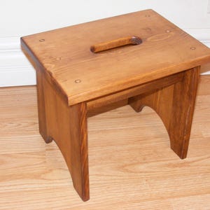 May include: A wooden step stool with a cutout handle in the top. The stool is stained a light brown color.