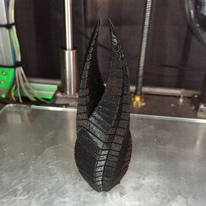 May include: A black, 3D-printed sculpture with a unique, geometric design. The object has a pointed top and a textured surface, likely created using a 3D printer. The sculpture is approximately 6 inches tall.