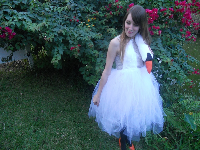 Pu&ograve; includere: Una donna che indossa un costume da cigno bianco con una gonna di tulle e un copricapo da cigno. &Egrave; in piedi davanti a un cespuglio verde con fiori rosa.