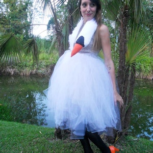 Pu&ograve; includere: Una donna con un abito di tulle bianco e scarpe arancioni con un accessorio per costumi a forma di testa di cigno. &Egrave; in piedi davanti a uno stagno con palme sullo sfondo.