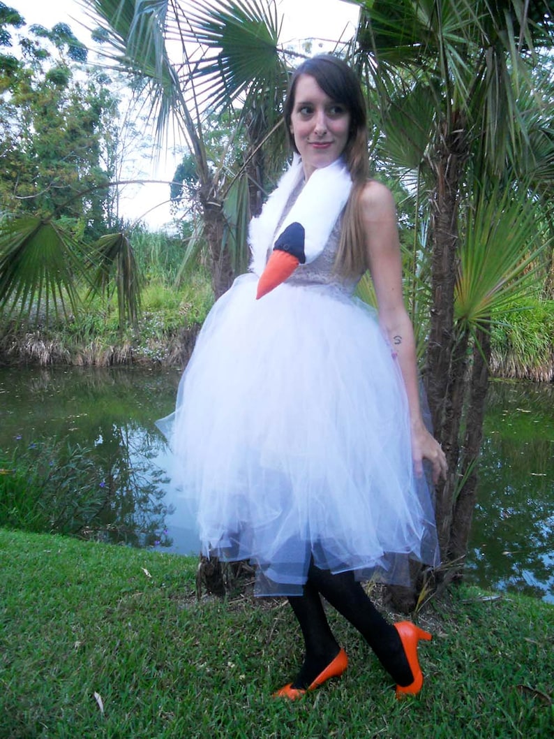 Pu&ograve; includere: Una donna con un abito di tulle bianco e scarpe arancioni con un accessorio per costumi a forma di testa di cigno. &Egrave; in piedi davanti a uno stagno con palme sullo sfondo.