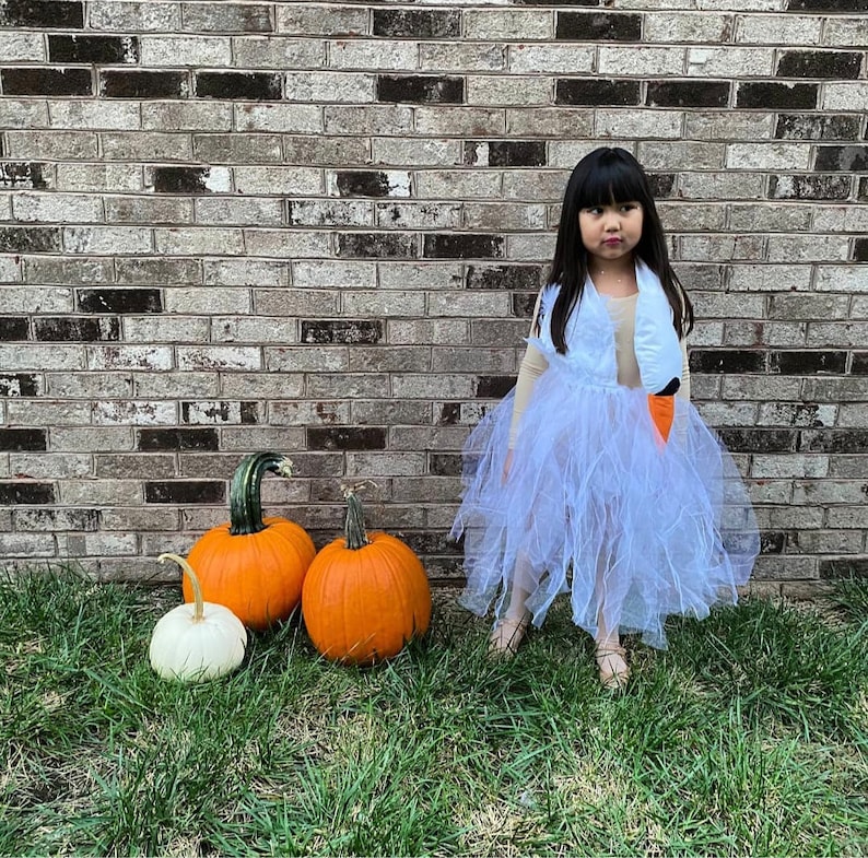 Pu&ograve; includere: Una bambina vestita da cigno per Halloween si trova davanti a un muro di mattoni. Indossa una gonna di tulle bianca e un costume da cigno marrone e bianco. Ci sono tre zucche davanti a lei, due arancioni e una bianca.