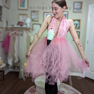 Pu&ograve; includere: Un tut&ugrave; di tulle rosa con una testa di fenicottero e piume attaccate. Il tut&ugrave; &egrave; indossato da una persona con leggings neri e piedi nudi. La persona &egrave; in piedi su un piedistallo bianco davanti a un tappeto rosa e bianco.