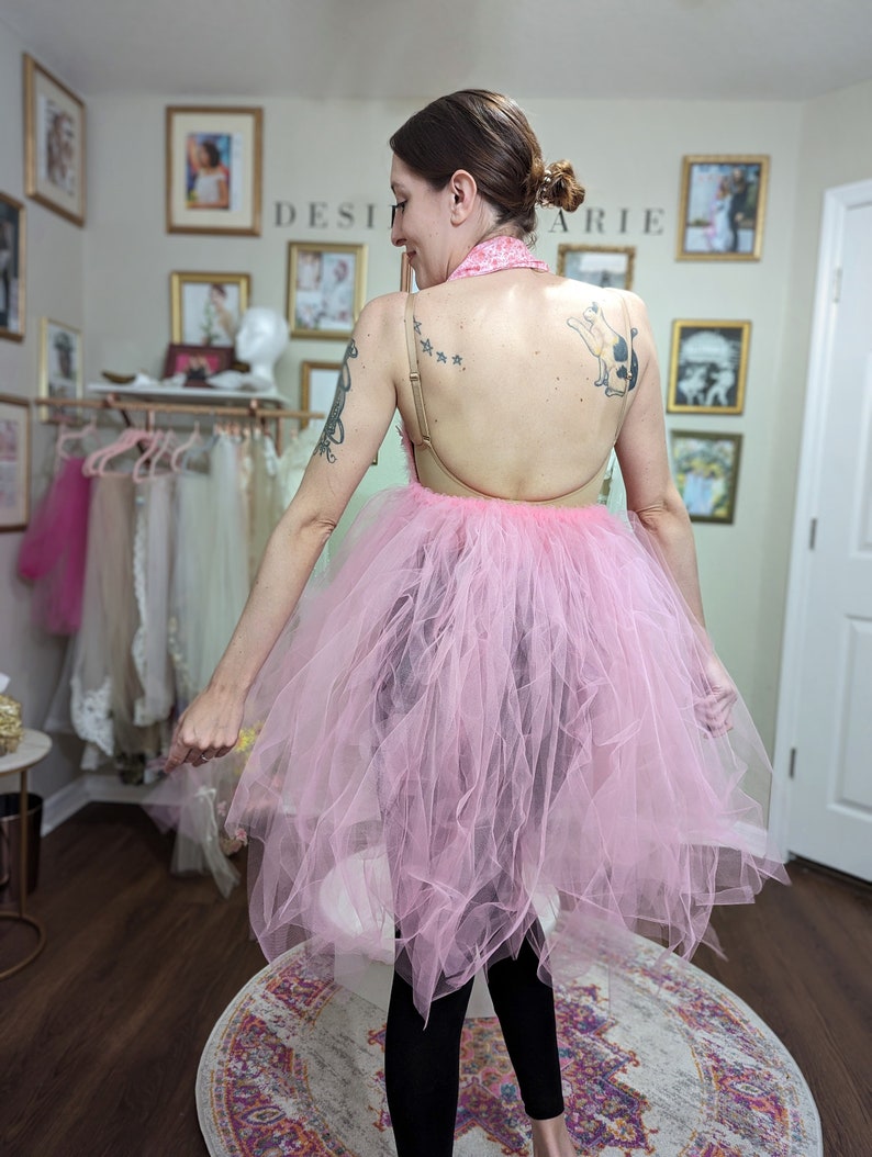 Pu&ograve; includere: Una donna indossa una gonna di tulle rosa con leggings neri. La gonna &egrave; molto ampia e ha molti strati. La donna &egrave; in piedi su un tappeto con un motivo floreale. Il testo "DESI" e "ARIE" &egrave; visibile sul muro dietro di lei.