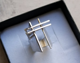 May include: A silver ring with a geometric design. The ring is in a black box with a white interior.