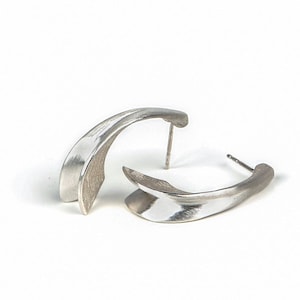 May include: A pair of silver stud earrings. Each earring features a curved, abstract shape with a smooth, reflective surface. The earrings are displayed on a white background.