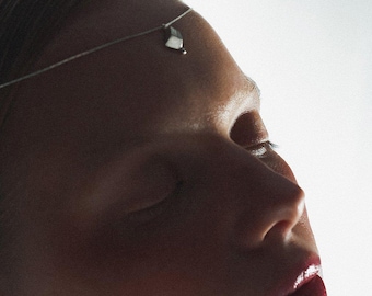 May include: A close-up of a person's face with closed eyes and a silver headpiece. The headpiece is a thin chain with a small, geometric pendant resting on the forehead. The lips are a deep red color.
