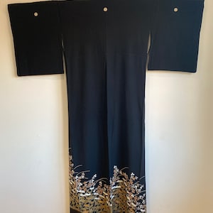 May include: Black kimono with floral design at the hem. The kimono has wide sleeves and is decorated with gold, white, and brown floral patterns. The garment is hanging against a white wall.