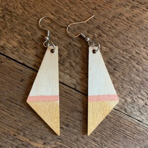 May include: A pair of wooden earrings with a geometric shape. The earrings are painted with a light pink and gold stripe.
