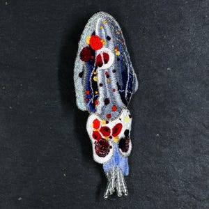 May include: A blue and white crocheted squid with red and yellow accents. The squid has a long, thin body with a rounded head and eight tentacles. The tentacles are decorated with beads and sequins.