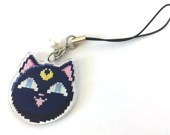 May include: A pixel art acrylic charm shaped like a cat head with dark blue fur, pink inner ears, light blue eyes, and a yellow crescent moon symbol on the forehead. The charm includes a silver metal jump ring, a small white bead, and a black cord.