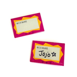 May include: Two rectangular name tag pins featuring a bright pink border with a jagged yellow inner frame. The center is a light gray rectangle with the text NICK NAME printed in black. One pin has the handwritten name Jojo and a star symbol.