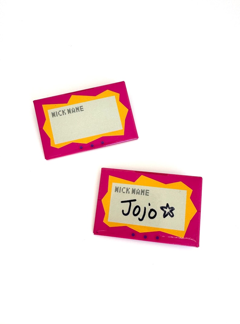 May include: Two rectangular name tag pins featuring a bright pink border with a jagged yellow inner frame. The center is a light gray rectangle with the text NICK NAME printed in black. One pin has the handwritten name Jojo and a star symbol.